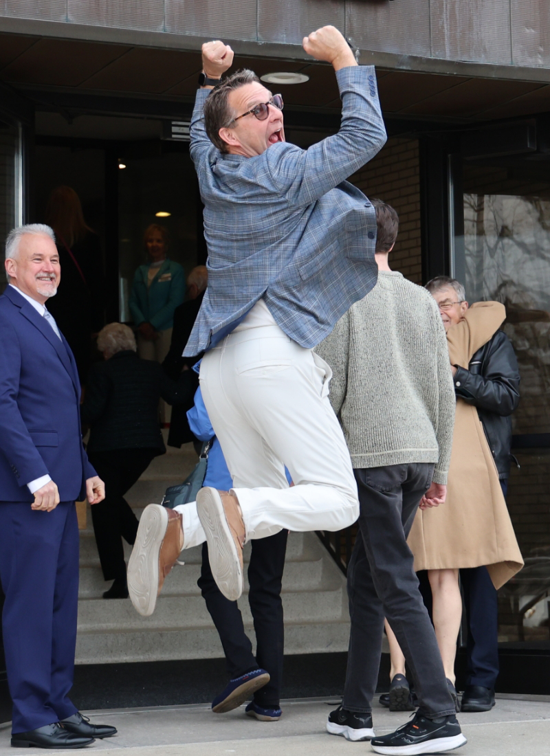 A man in a blazer and white pants jumps enthusiastically outside a Local Church in Woodbury, while others stand and interact in the background.