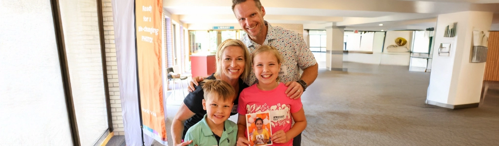 A smiling family of four poses indoors; the girl in front holds a book while the others stand closely behind her, serving warmth and togetherness.