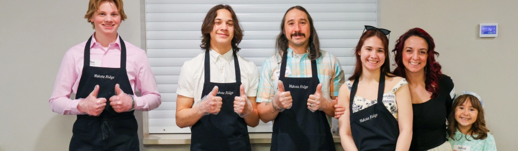 Six people, some wearing aprons, stand side by side indoors and smile at the camera; two give thumbs up, proud of serving together.