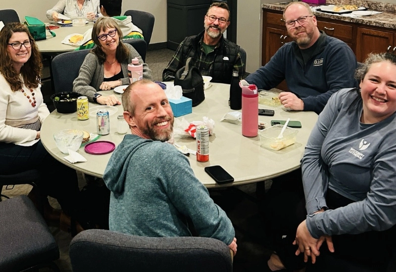 Six adults, each from diverse careers, sit around a round table with food and drinks, smiling at the camera in an indoor setting.