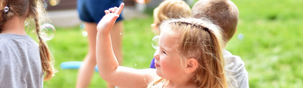 Children in Preschool Programs play outside on grass, with one girl reaching up towards floating soap bubbles.