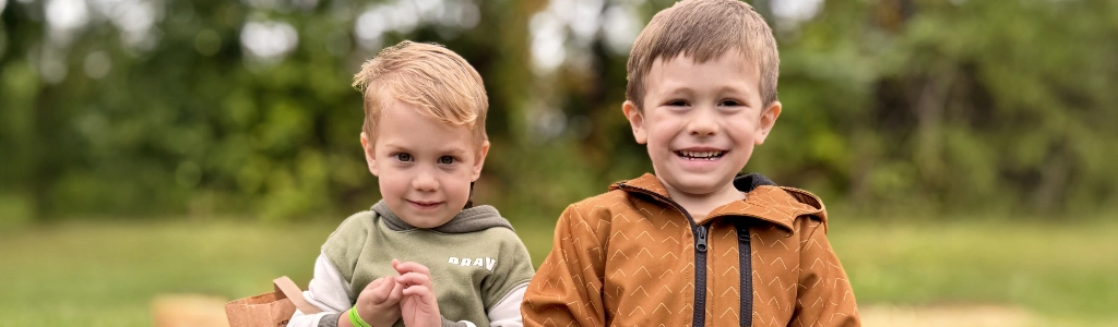 Two young boys sit side by side outdoors on a blanket, smiling at the camera with green trees blurred in the background—capturing the joy of friendship often found in Preschool Programs.