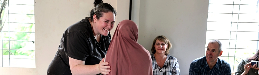 A woman smiles and interacts with a girl in a brown hijab while two adults sit and watch in a bright room with barred windows, discussing possible Next Steps.